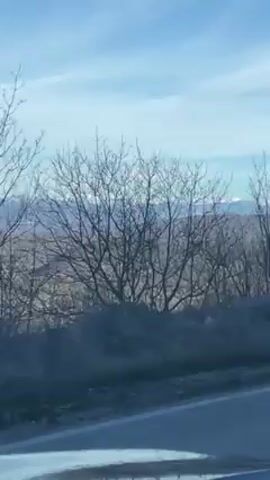 The video shows a view from a car driving on a road. In the distance, a mountain range with snow-capped peaks is visible under a blue sky with scattered clouds. The foreground features a landscape of low-lying hills and sparse vegetation, possibly burnt trees or shrubs. A roadside barrier is briefly visible next to the road.