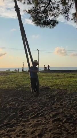 A child wearing a hat and blue shirt is swinging on a tire swing attached to a tree. The swing is made of two thick ropes. The child is positioned in the center of the frame, in motion. The background shows a grassy area with some trees, power lines, and a body of water, likely the sea, with a cloudy sky. Several other people are visible in the distance near the water. The ground in the foreground is sandy or dirt. The sun is setting, casting a warm glow.