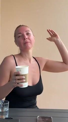 In the video, a woman with light-colored hair pulled back is seated at a dark table outdoors, possibly on a balcony or patio, against a plain light-colored wall. She is wearing a black spaghetti-strap top. In her left hand, she holds a white disposable cup, which appears to be from Starbucks, given the visible green logo. A clear glass with water and a smartphone are also visible on the table.

The woman is actively singing along to music, with her mouth wide open and her head tilted back, eyes often closed, conveying an emotional and expressive performance. Her right hand moves dramatically with the music, at times raised and open, emphasizing her singing. She appears to be enjoying herself thoroughly, embodying the song. A ring is visible on her left ring finger, and her nails are manicured. The scene is brightly lit, suggesting it is daytime.