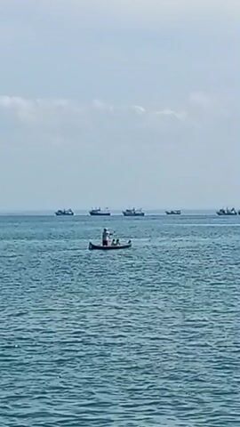 A wide shot captures a serene ocean scene under a hazy sky. In the foreground, gentle waves lap against an unseen shore. The midground is dominated by a small canoe, where a person stands, appearing to cast something into the water. Another person is seated in the canoe. Further in the distance, a line of larger ships, likely fishing vessels, are spread across the horizon, their silhouettes distinct against the pale sky. The overall atmosphere is one of tranquility and perhaps a hint of industry with the presence of the fishing fleet.