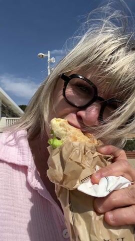 A woman with blonde hair and glasses eats a sandwich on a sunny day at the beach. She wears a pink shirt and holds the sandwich, which appears to contain bread, lettuce, and other ingredients, in a brown paper bag. A white cloth napkin is also visible in her hand. The background shows a beach with sand, some buildings, a streetlight, and a blue sky with some clouds. Her hair is windswept across her face. The focus of the shot is on the woman and the sandwich she is eating.