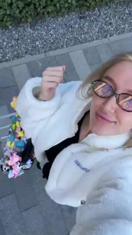 The video shows a person with blonde hair, wearing oval glasses with a tortoiseshell pattern, a white, fluffy jacket, and a black shirt underneath. They are standing on a paved sidewalk with gray stones on one side and dark gray bricks on the other. The person is holding up their fist, as if offering a fist bump. Next to them is a colorful assortment of small, plastic toys, possibly animal figures. The video appears to be taken at a slightly low angle, looking up at the person.