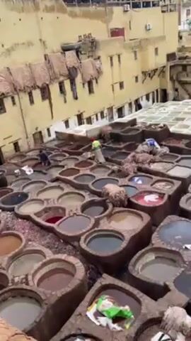 The video shows an aerial view of a tannery in what appears to be an older city. The tannery consists of numerous stone or concrete vats filled with liquids of various colors, including red, brown, white, and gray. Several people are working in the vats, some standing and moving around, and others kneeling or bent over. The vats are tightly packed together. Buildings surround the tannery on multiple sides, some with clotheslines strung up with drying items. The buildings are mostly tan or light brown in color.  The overall scene depicts a traditional method of leather tanning in a historical setting.