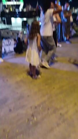 The video shows a group of people dancing on a wooden boardwalk at night. A young girl in a white dress is walking across the boardwalk, while other people are dancing and socializing in the background. There is a boat docked nearby, with lights and people on its deck. The overall atmosphere appears to be festive.