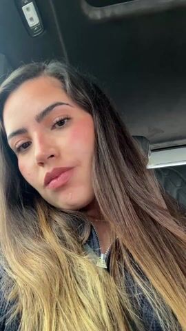 A woman is sitting in a car, looking at the camera. She is wearing a dark blue zippered sweater and a gold necklace. She has long brown and blonde hair. The car has a black interior. The woman makes a face for the camera. In the background, music is playing.