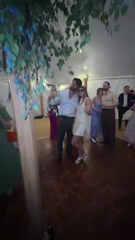 The video appears to be filmed at a wedding reception or similar event held in a tent with a wooden floor and ceiling decor of hanging leaves and lights. The first few seconds show a couple in formal attire standing near a wooden pillar, looking up. The video then transitions to a more dynamic scene with a band playing on a stage illuminated by blue and purple lights. People are visible dancing and socializing. The band members are playing instruments, and some attendees are dancing. The scene shifts back to the couple and other guests. One shot shows a man in a suit and tie holding a bottle. The video ends with a brief shot of the couple from the beginning, now with other guests in the background. The overall atmosphere seems festive and celebratory.