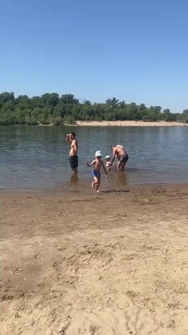 A group of people are at the edge of a body of water. The water appears to be a river or lake. The foreground is a sandy beach. In the background, there are trees and a clear blue sky. One person is standing in the water on the left side of the frame, facing away from the camera. In the center of the frame, two people are interacting in the shallow water. One person is carrying another person on their back. Further to the right, a child is running on the sandy beach towards the water. The lighting suggests it is daytime.