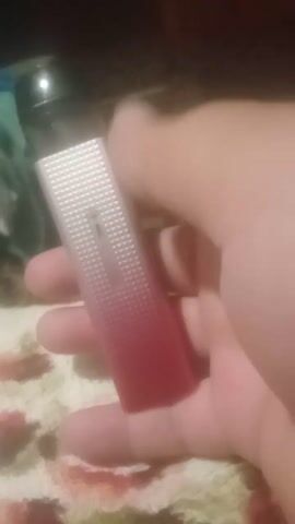 The media file is a close-up, handheld video of a person's hand holding a small, rectangular object that appears to be a vape device. The object has a silver, ribbed section at one end and a red, gradient section at the other. The lighting is dim, suggesting an indoor setting, possibly in the evening. The background is out of focus but shows a patterned fabric, likely a blanket or towel. There is no discernible text visible on the object or in the scene. The activity appears to be simply holding and displaying the vape device. The image quality is somewhat blurry, making it difficult to discern specific details about the person's hand or the exact surroundings. The location context provided is Pavlivka, Ukraine, but this information cannot be verified from the visual content alone.