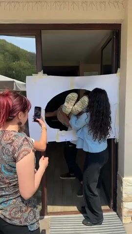 In Malka Vereya, Bulgaria, a daytime scene unfolds at a doorway with natural light indicating sunny weather. A woman with long dark hair, wearing a light blue denim-like shirt and dark pants, is engaged in a playful activity. She is seen pulling a man, dressed in a white t-shirt and light brown pants, horizontally through a circular hole cut into a large white sheet that is taped to the door frame. Her light green and white chunky sneakers are prominent as she struggles and laughs. Another man with dark hair and a beard is visible behind the sheet, also laughing, seemingly assisting or participating. A fourth person, mostly out of frame, is using a smartphone to record the event. All participants are smiling and laughing exuberantly, suggesting a fun and lighthearted game. No discernible text is visible on any objects or clothing in the frame.