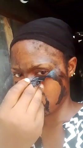 A young woman with dark skin applies a dark substance to her face from a small square of black material. She wears a black head covering and a black and white patterned top. She makes a kissing face for the camera. She is indoors with a dark doorway behind her.