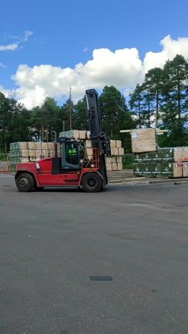 This is a video clip showing a red forklift truck in operation at an outdoor lumber yard. The forklift has a black carriage and lift mast, and its cabin is occupied by a person wearing a green safety vest and a helmet. The truck is positioned on a paved surface, with stacks of lumber visible in the background. Some of the lumber is wrapped in dark green plastic with white lettering that reads "WOOD TERMOO".

The scene is set against a backdrop of tall green trees and a bright blue sky with white, fluffy clouds. The lighting suggests it is daytime, likely mid-morning or afternoon, with clear weather. The forklift appears to be engaged in moving or stacking lumber, as its mast is raised and positioned near the stacks. The overall atmosphere is one of industrial activity in a natural, wooded environment. No specific emotions are discernible, but the presence of safety gear suggests a professional work setting. The image is oriented vertically in the video.