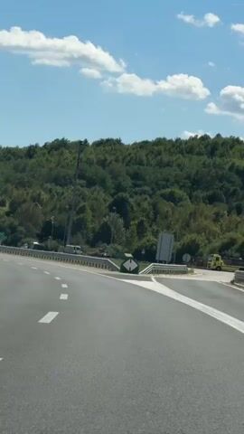 A paved road with white lane markings curves to the right. Tall streetlights stand along the side of the road. A billboard with colorful graphics is visible on a hillside covered in green trees and foliage. Several cars are on the road, and a guardrail lines the edge of the pavement. The sky is blue with scattered white clouds.