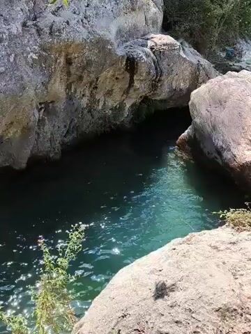 The video shows a group of people swimming and relaxing in a natural, clear blue body of water surrounded by rocks and lush green vegetation. The scene appears to be a popular outdoor recreational spot, possibly a river or a natural pool. The water is calm and inviting, with several individuals enjoying a swim. The overall atmosphere is one of leisure and enjoyment of nature.