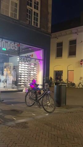 A medium shot shows a street scene at night. In front of a brightly lit storefront of a shoe store, a person dressed in a green suit with a matching mask sits on the ground playing a guitar. A black bicycle is parked in front of the person. Other pedestrians walk by. To the right of the shoe store, there is a yellow building with a few parked bicycles. A black trash can sits in the foreground. The street is paved with brick. The lighting changes behind the shoe store window.