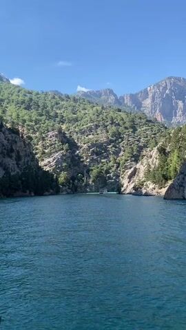 The video captures a scenic view of a wide body of water, likely a lake or reservoir, nestled between towering mountains. The water in the immediate foreground is a deep blue with gentle ripples, suggesting the camera is on a moving boat. Further into the distance, the water transitions to a vivid turquoise hue. A white, multi-decked passenger boat is visible on this turquoise stretch of water, appearing to be stationary or moving slowly. To the left of the large boat, a smaller, dark-colored vessel, possibly a kayak or a small paddle boat, is also present on the water. The surrounding mountains are steep, covered extensively with lush green trees and shrubs, interspersed with exposed grey rock formations. The sky above is clear and blue with a few wispy clouds, indicating bright, sunny daytime conditions. Indistinct human voices can be heard in the background. The location context provided is Akseki, Turkey.