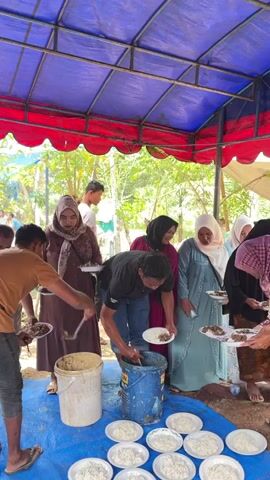 The video shows a group of people, mostly women wearing hijabs, standing in a line under a blue and red tent, being served food. Two men are serving food from large buckets into plates held by the people in line. One man is wearing a brown shirt and the other is wearing a black t-shirt and jeans. Several white plates are on the ground in front of the serving area, some filled with rice. The environment appears to be outdoors, with trees visible in the background. The overall scene suggests a communal meal or a catering event.