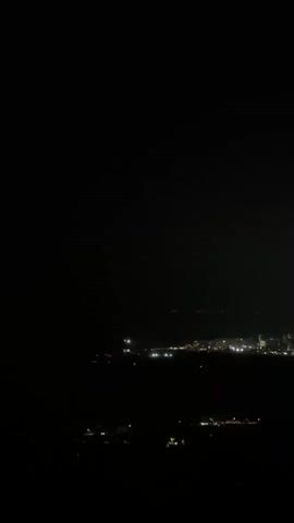The video displays a dark outdoor scene at night, with a city skyline visible in the distance. Lights from buildings illuminate the cityscape, and a few faint lights suggest activity in the foreground. The overall impression is of a city viewed from a slightly elevated, distant vantage point.