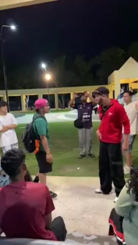 A group of people are gathered in an outdoor public area at night. Artificial lights illuminate the scene. The focus is on two men facing each other in a small open space. One man is wearing a red hooded sweatshirt and black pants, with a cap and sunglasses. The other man is wearing a green t-shirt, black shorts, a pink hat, and is carrying a backpack. They are gesturing and appear to be engaged in some form of competitive verbal exchange, possibly a rap battle or freestyle. Around them, other individuals are watching, some recording the event with their phones. The background shows green grass, tiled walking areas, and a low building with open archways and windows. Some figures are sitting on a tiled surface in the foreground. The overall atmosphere suggests an informal, impromptu gathering centered around a performance.