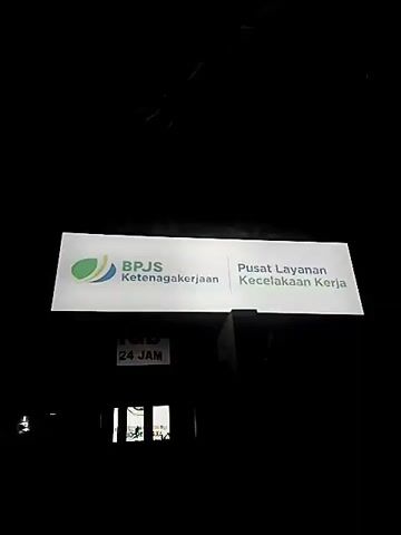 The video shows a lighted sign for BPJS Ketenagakerjaan, which translates to "Employment Social Security Agency". The sign also says "Pusat Layanan Kecelakaan Kerja", meaning "Work Accident Service Center". Below the main sign, a smaller sign indicates "24 JAM", meaning "24 Hours". The background is dark, suggesting it is nighttime. There are some blurred elements in the foreground and background, possibly trees or other foliage, and some faint reflections. The overall impression is of an outdoor scene at night, with the illuminated sign being the primary focus.