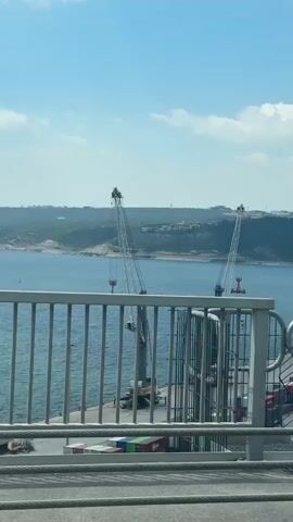 The video shows a view from a high vantage point, likely a bridge, looking out over a harbor or port. Two large cranes stand on a pier, ready to load or unload cargo. Stacks of colorful shipping containers are lined up along the pier. Beyond the pier, there is a body of water and a hilly coastline. The sky is blue with scattered clouds. The view is partially obscured by a metal railing in the foreground. The camera appears to be moving.