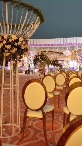 This video captures an elegantly decorated event space, likely for a wedding or formal reception, in Askihala, India. The foreground shows multiple rows of empty chairs with gold frames and cream-colored cushions, arranged facing a stage or main area.

To the left, a large decorative structure made of white poles holds a substantial floral arrangement with white, yellow, and purple flowers. Similar large floral displays and decorative elements are visible on and around the stage area in the background. The stage itself is elaborately decorated with numerous flowers, lights, and shimmering elements, creating a lavish and festive atmosphere.

The lighting suggests it is evening or night, with artificial lights illuminating the decor. A dark, possibly tented, ceiling covers the event space. The overall scene indicates preparation for a grand event, with no guests present yet. No visible text is present in the video.