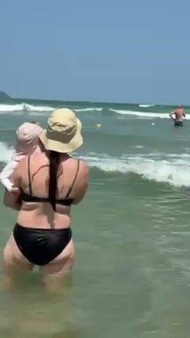 The video depicts a beach scene on a sunny day. A woman in a black bikini and a tan hat is carrying a toddler who is wearing a pink hat and a light-colored outfit, wading into the ocean. Other individuals are visible further out in the water, with waves breaking in the background. The water appears green and slightly choppy. The sky is a clear blue.