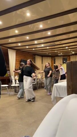 The video captures a lively indoor gathering, showcasing a group of approximately ten young adults, predominantly women, engaged in social dancing. They are dressed casually in t-shirts (black and white being dominant colors), loose-fitting jeans, sweatpants, and comfortable footwear like sneakers and purple Crocs. Their expressions range from focused to joyful, with many smiling and laughing as they move in unison, possibly performing a line dance or choreographed steps.

The setting is a spacious room with warm, inviting decor. The walls and ceiling are paneled with light-colored wood, featuring dark wooden beams across the ceiling from which numerous recessed spotlights cast a bright, warm glow. Several rectangular tables, draped with white tablecloths and surrounded by white chairs, are arranged around the perimeter, suggesting a dining or reception area. Some bottles and glasses are visible on the tables, implying refreshments are available. A large, dark-screened television stands on a low table against one wall, and white curtains adorn the windows. Subtle fairy lights add to the festive ambiance in the background. The floor is a light, neutral color.

The overall atmosphere is energetic and celebratory, indicative of a social event or party, likely taking place in the evening or night given the reliance on artificial lighting. No distinct, legible text is visible on clothing or signage. The location is Yus'ki, Russia.