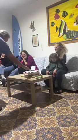 Here is a detailed description of the video:

The scene shows a group of people gathered in a living room, presumably celebrating a birthday.  A man is seen lighting candles on a cake placed on a small coffee table. The cake appears to be decorated with frosting and some dark-colored toppings. A woman holding a young child on her lap leans forward to blow out the candles. Other people sit on a couch nearby watching. The room is decorated with a large painting of fish and a surfboard leaning against the wall. The floor is covered with patterned ceramic tiles.
