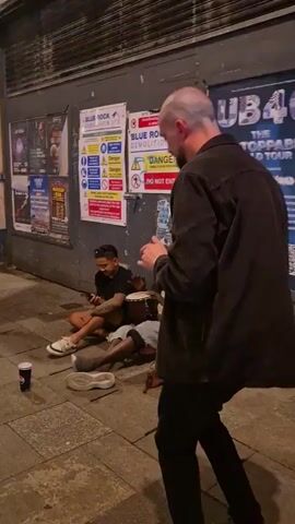 The video shows three men outside a building that has a rolled-down door. One man is sitting on the ground looking at his phone. Another man is sitting beside him, holding a drum. A third man standing looks on with his hands moving as if he is playing the bongos. The building has posters and signs on the wall, including one for Blue Rock Demolition Ltd. and one for UB40's "Unstoppable World Tour". There is a cup on the ground in front of the man who is standing.