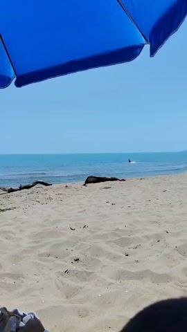 A person is sitting under a blue umbrella on a sandy beach. The ocean is visible in the background with a clear blue sky. There is a piece of driftwood on the sand near the water's edge. In the distance, a person can be seen in the water. A small drink with a lime is on a tray in the foreground.