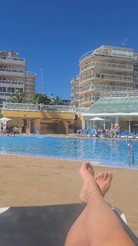The video shows a swimming pool on a sunny day. 
Two multi-story buildings flank the pool, each with white exteriors and balconies. The pool area is crowded with people swimming and lounging on deck chairs, shaded by white umbrellas. Palm trees are scattered around the pool area.
The video begins with a view of a person's legs resting on a lounge chair in the foreground, feet crossed. The camera pans slowly to the left, showing more of the pool and the surrounding buildings. A small waterfall feature can be seen in the middle of the pool. The sky is clear and blue.