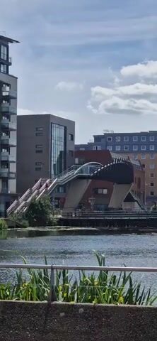 The video shows a cityscape by a body of water, possibly a canal or river.  There's a distinctive modern bridge with a covered walkway that appears to connect two buildings. Several buildings of varying heights and architectural styles are visible in the background.  The sky is blue with scattered white clouds.  The camera pans upward to show an airplane flying across the sky.  Two people walk in front of the camera. The railing of the waterfront is in the foreground along with some foliage. The water appears calm.