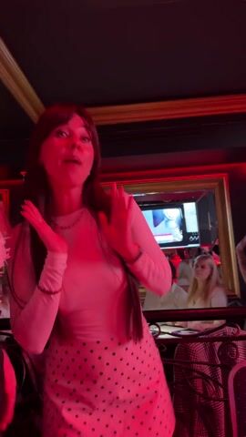 A woman in a white long-sleeved shirt and black polka dot skirt dances while lip-syncing in what appears to be a restaurant or bar. She has long brown hair with bangs and is wearing jewelry. Behind her, a mirror reflects other people seated at tables. The lighting is dim, with a red light strip above. A television screen in the background shows what appears to be a male news anchor. The video appears to be recorded on a mobile phone.
