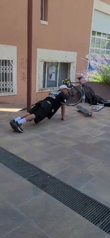 A person in a white cap and dark clothing is doing push-ups on a paved area. Another person in a white tank top and dark pants walks into the frame from the right. In the background, there is a building with a pinkish-orange facade, a window with a metal grate, and a bicycle. There are also skateboards visible on the ground.