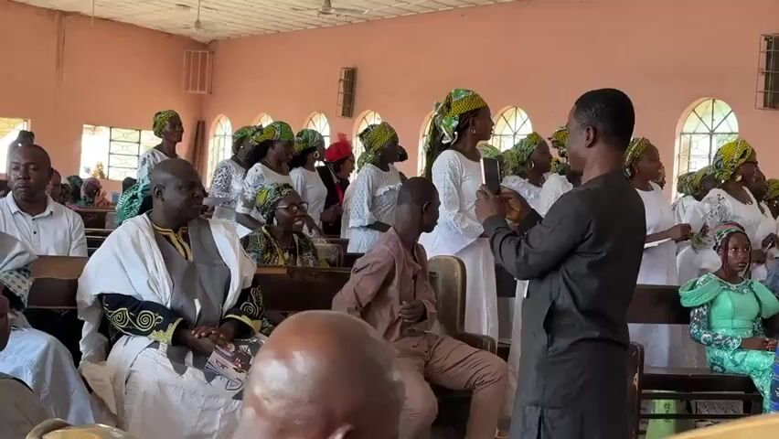 A group of people are gathered indoors, appearing to be in a church or hall. Many are wearing white clothing and headscarves, suggesting a uniform or celebratory attire. A man in the foreground on the right is holding up a smartphone, seemingly recording or taking photos of a woman in white who is facing him. This woman and others around her are looking towards the man with the phone and appear to be singing or engaged in a lively activity. Some people are seated on wooden benches, while others are standing. The walls of the room are painted a light pink, and there are windows with arched tops. Decorations, possibly streamers or garland, are visible near the ceiling. The overall atmosphere seems festive and communal.