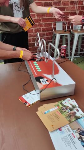 The video shows a close-up of a game involving a metal maze, played by several people. The game is set on a brown wooden table, with a brick wall in the background.

**Subjects:**
* **People:** The hands and arms of at least three people are visible, actively participating in the game. One person is wearing a yellow wristband.
* **Objects:** The central object is a silver and white game console with a red "Doppel Herz" logo and several green lights. A can of Coca-Cola is perched on top of the maze. There are also some promotional flyers and brochures on the table.
* **Scene:** The scene appears to be an outdoor event or exhibition, possibly a fair or a promotional stand, given the presence of the game and promotional materials.

**Activity or Event:**
The people are engaged in playing the maze game, with one person holding a tool that seems to guide a metal ball through the maze. The game console has lights that might indicate progress or scoring. The Coca-Cola can appears to be part of the game's setup or a prize.

**Notable Details:**
* **Time of Day/Weather:** The lighting suggests it could be daytime, possibly outdoors.
* **Location Cues:** The brick wall and outdoor setting hint at a specific location. The promotional materials are in Russian, which aligns with the provided location context of Il'inskoe, Russia.
* **Emotions:** The focus on the hands and the game suggests concentration and engagement from the players.

**Visible Text:**
* "Doppel Herz" (logo)
* Russian text on the promotional flyers and the game console, though it's difficult to read clearly. One flyer has text that appears to be related to vitamins and health supplements, possibly mentioning "Витамин Q10" and "В-Комплекс."
