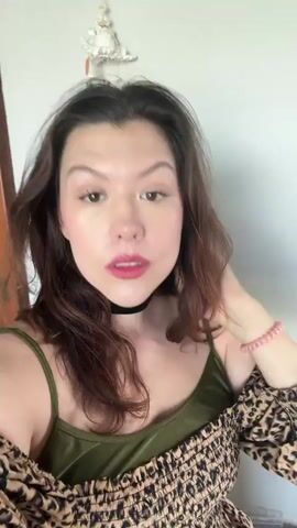 A woman with long brown hair, wearing a green top and a leopard print overlay, is looking at the camera and touching her neck. She has on red lipstick and a black choker necklace. There is a light-colored wall behind her, and a decorative item hanging on the wall to her left.