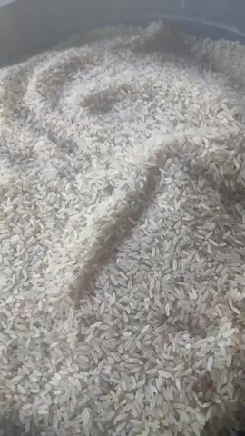 The media file shows a close-up, overhead view of a large quantity of uncooked white rice in a dark container. The rice is piled high, with visible ripples and contours created by the grain. The lighting suggests it is daytime, though the specific time and weather conditions are not discernible. There are no people, animals, or discernible text in the frame. The scene is static, with no apparent activity taking place, and the focus is entirely on the texture and volume of the rice. The setting appears to be indoors or a sheltered outdoor space, given the lack of environmental elements like wind or strong sunlight. The overall impression is one of abundance or preparation for a large meal.