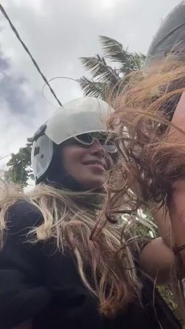 A woman wearing a helmet and a black t-shirt rides on a motorcycle. Her blonde hair blows in the wind, obscuring parts of her face. The video captures her from the chest up, with a cloudy sky and glimpses of trees in the background. The camera appears to be mounted on the motorcycle, giving a first-person perspective.