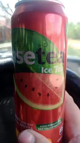 A person's thumb and forefinger hold a red soda can featuring a label with a slice of watermelon and the text "fusetra Ice Tea". The background is blurred, suggesting the can is inside a vehicle.