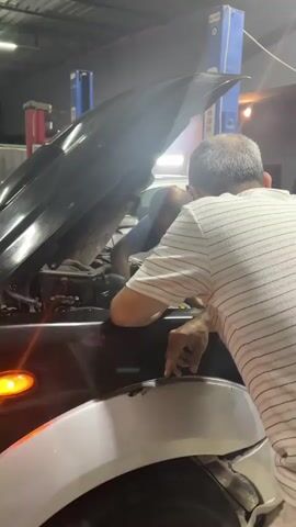 Two men are working on the engine of a black car in a garage. The car's hood is open. One man, wearing a blue shirt, is leaning into the engine compartment, while the other, wearing a white and gray striped shirt, is pointing at a spot on the car's body near the front wheel. The garage has a car lift in the background. The lighting is bright, and the scene appears to be focused on diagnosing or repairing the car.