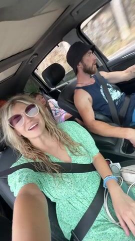 The video shows four people in a car. In the front passenger seat, a woman with blonde hair and sunglasses is talking to the camera, wearing a green floral dress. To her right, a man with a beard and a black hat is driving, wearing a blue tank top and blue shorts. In the back seat, a woman with short red hair and glasses is in the middle, wearing a colorful floral dress. Next to her is a young girl with brown hair, wearing a pink and white tie-dye hoodie. The car appears to be moving along a road with vegetation visible outside the windows.