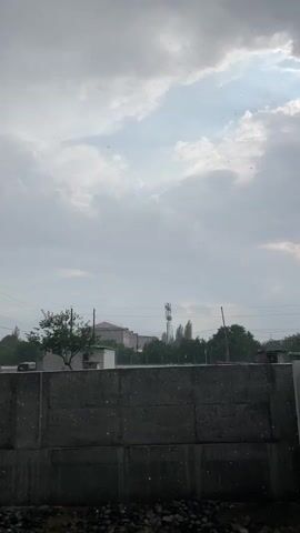 The video shows a view of a cloudy sky with a concrete wall in the foreground. Behind the wall, there are trees, buildings, and a communication tower. Small white particles are visible in the air, suggesting it might be raining or snowing. The overall atmosphere appears overcast and potentially inclement.