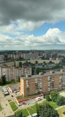 The video shows an aerial view of a city with multiple residential buildings and green spaces under a cloudy sky. In the foreground, a large orange brick apartment building with many balconies is prominent. To its left is a smaller building with a red roof, possibly a commercial space, and a parking lot with a few cars. Further into the background, there are rows of apartment buildings of varying heights and architectural styles, some older and some newer. Trees and green areas are interspersed between the buildings. The distant skyline features more buildings and a tall antenna structure. The sky is a mix of blue patches and white, fluffy clouds, suggesting daytime.