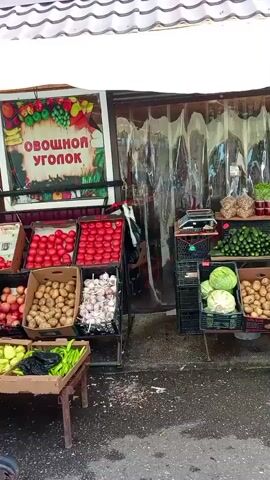 A short, vertical video shows an outdoor fruit and vegetable stand. The stand has a sign that reads "ОВОЩНОЙ УГОЛОК", which translates to "Vegetable Corner." Various produce, including tomatoes, potatoes, garlic, cucumbers, cabbage, and peppers, are displayed in crates and baskets. There are also jars of pickles and some bagged nuts on display. To the right of the produce display, a large barrel with a sign reading "КВАС" (Kvas) is visible. The ground appears wet, suggesting it may have rained recently. The overall impression is of a small, local market stall.