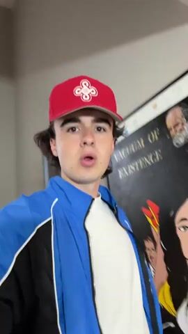 A young person wearing a red baseball cap and a blue and black jacket is speaking to the camera. They are in a room with artwork on the walls. The artwork includes a portrait of an old man with a white beard and a painting of two women. The person is gesturing with their hands as they speak. The video appears to be a vlog or a personal update.