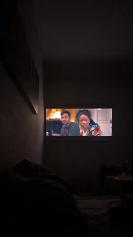 A television screen displays a scene from a show or movie. On the left side of the screen, a man with dark hair and a beard, wearing a dark sweater, is looking to his right. On the right side of the screen, a woman with dark, curly hair, wearing a colorful, patterned jacket and a white shirt, is looking directly ahead with a concerned expression. The background behind them is blurred, but appears to be an indoor setting. The television is mounted on a dark wall, and a power cord is visible hanging down from the bottom of the screen. The lower portion of the image is very dark, with the suggestion of a bed or couch covered with blankets in the foreground. To the right of the television, on a table or surface, are some indistinct objects, possibly a bottle and other items. The overall scene is dimly lit, with the light from the television screen illuminating the immediate area.