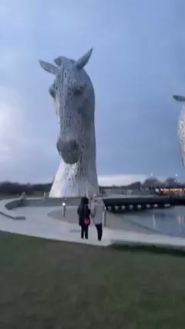 Two people stand on a paved walkway near a body of water with two enormous horse head sculptures looming over them. The sculptures are made of a patchwork of metallic plates and appear to rise out of the water. The sky is overcast with gray clouds. The walkway is bordered by a patch of green grass.