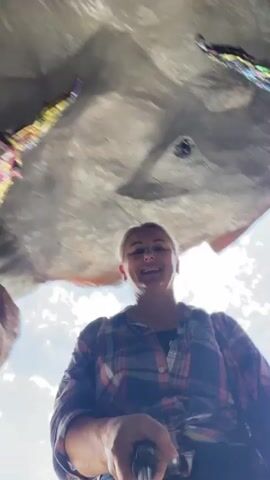 A low-angle, first-person perspective video shows a woman with blonde hair pulled back, wearing a red, blue, and white plaid shirt, holding a selfie stick. The woman is smiling and looking up at the camera. The background appears to be a cave-like structure with textured grey walls and some colorful decorations. The woman's right arm is extended outwards, gesturing towards the right side of the frame. The video is blurry in places due to movement.