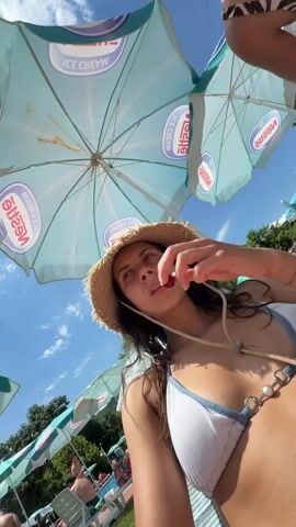 A low-angle, wide shot shows a woman in a white bikini and a straw hat from the chest up, looking at the camera. She is under two blue and white umbrellas, each with the Nestle Ice Cream logo printed on it. Her left hand is raised to her mouth, and she appears to be eating something. The background is blurred, but appears to be a crowded outdoor setting, possibly a beach or pool area, with other umbrellas and people visible. The sky is visible above the umbrellas and is partly cloudy.